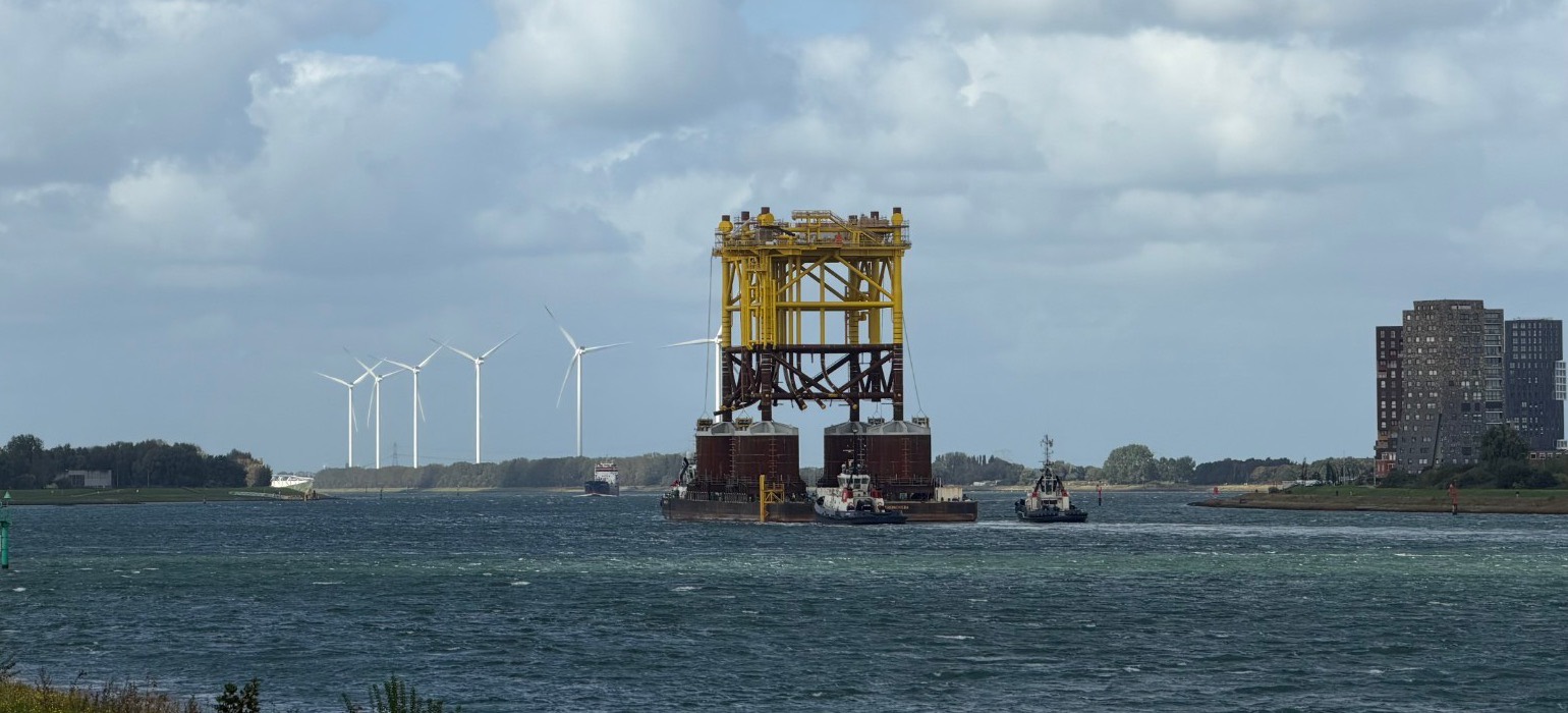 Yellow offshore structure on a pontoon, escorted by tugs, with wind turbines in the background.